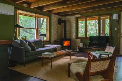 Cozy living room with a gray sofa, wood stove, large windows, and natural decor, surrounded by greenery.
