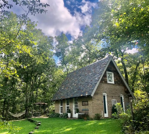 A charming brick cottage with a slate roof, surrounded by trees and a green lawn, featuring a hammock in the background.