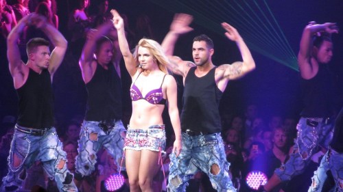 A performer in a sparkly outfit dances on stage with backup dancers, surrounded by colorful lights and an enthusiastic crowd.
