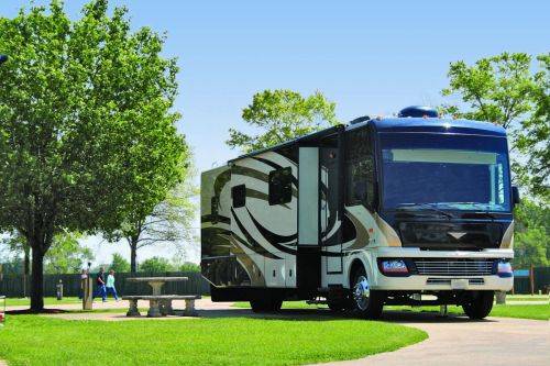A large RV parked on a grassy area with trees and a clear blue sky in the background.