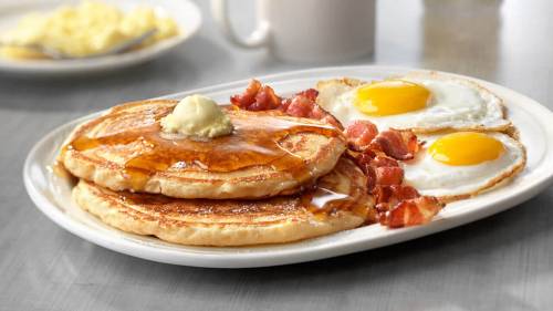 A plate of pancakes topped with butter and syrup, served with crispy bacon and two sunny-side-up eggs.