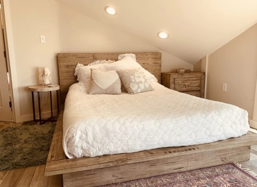 Cozy bedroom with a wooden bed, decorative pillows, and a small nightstand in a softly lit, sloped ceiling space.