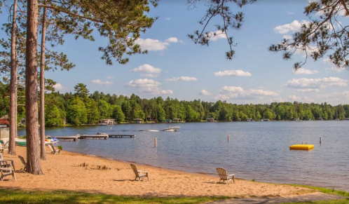 A serene lakeside scene with sandy beach, boats, and lush green trees under a blue sky with fluffy clouds.