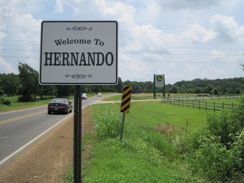 Welcome sign for Hernando beside a road, with green fields and a cloudy sky in the background.