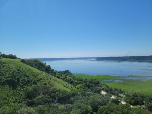 A scenic view of a calm lake surrounded by green hills and trees under a clear blue sky.