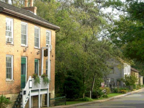 A quaint brick building with green doors and a white staircase, surrounded by trees and a quiet street.