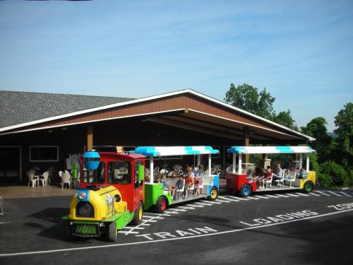 Colorful train-shaped ride vehicles are parked outside a building with a covered area and people seated at tables.