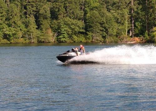 A person rides a jet ski on a lake, creating a spray of water, surrounded by lush green trees.