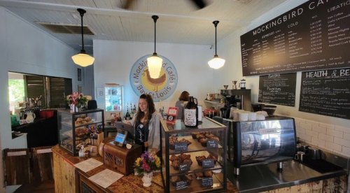 A cozy café interior with a smiling barista at the counter, displaying baked goods and a menu on the wall.