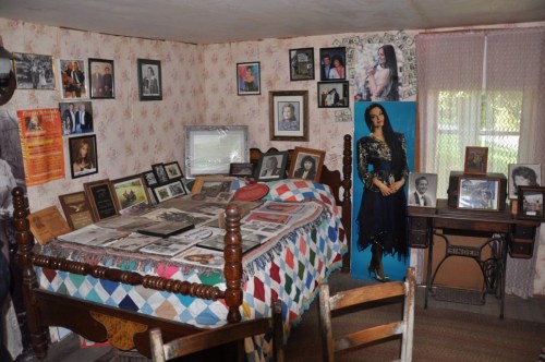 A cozy room with a bed covered in quilts, walls adorned with framed photos, and a life-sized figure in a blue dress.