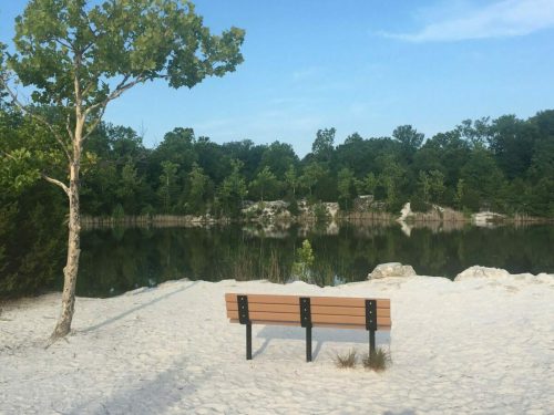 A wooden bench on a sandy shore overlooking a calm, reflective lake surrounded by trees.