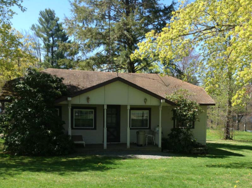 A small, charming house surrounded by greenery and trees, with a front porch and a well-maintained lawn.