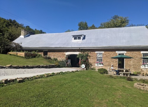 A rustic stone building with a metal roof, surrounded by greenery and a gravel path, under a clear blue sky.