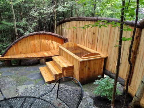 A wooden hot tub and seating area surrounded by trees and a rustic wooden fence.