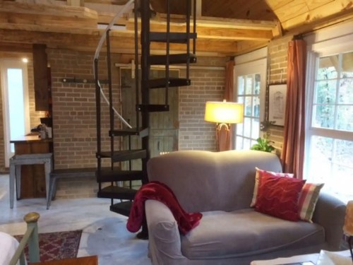 Cozy interior of a cabin featuring a spiral staircase, a sofa with a red throw, and large windows.