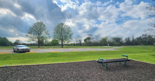 A grassy area with a bench, trees, and a parked car under a partly cloudy sky.
