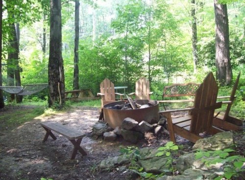 A cozy campsite with a fire pit surrounded by wooden chairs, a hammock, and lush green trees.