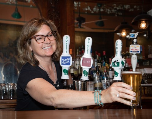 A smiling bartender serves a pint of beer from a tap with various beer handles in a lively bar setting.
