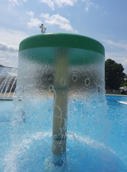 A circular water feature with a green top, spraying water into a blue pool under a clear sky.