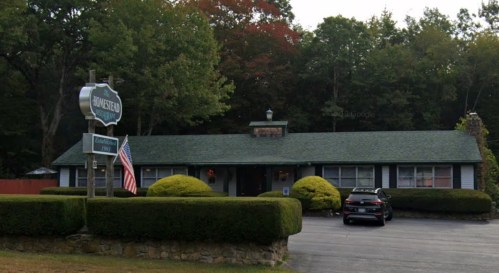 A cozy, single-story building with a green roof, surrounded by hedges and trees, featuring a sign that reads "Homestead."