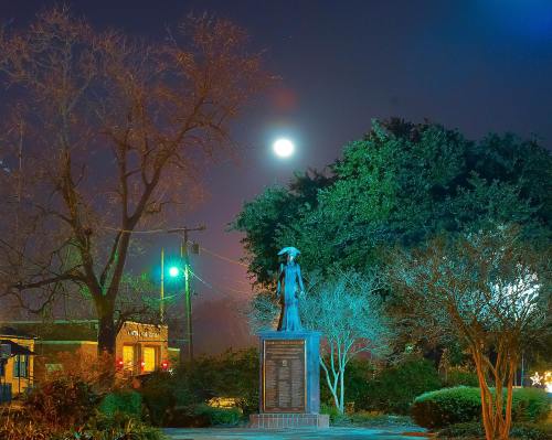A statue stands in a park under a bright full moon, surrounded by trees and a softly lit night sky.