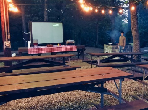 A dimly lit outdoor area with picnic tables, a projector screen, and a person standing near a grill. String lights overhead.