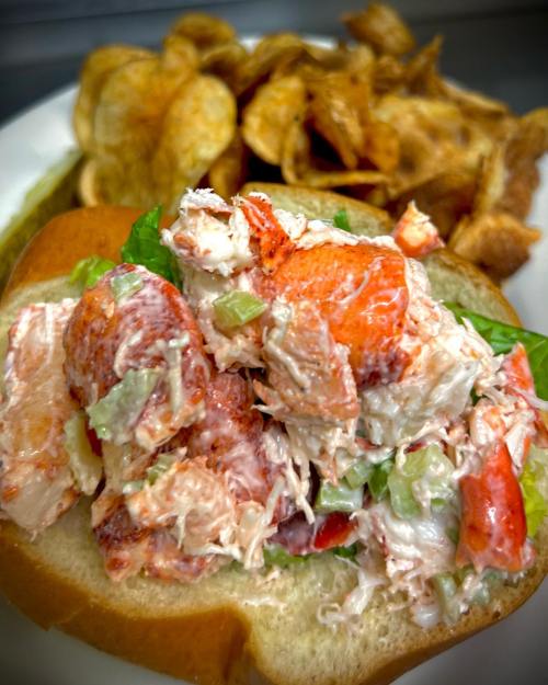 A lobster roll filled with lobster salad on a bun, served with a side of crispy potato chips.