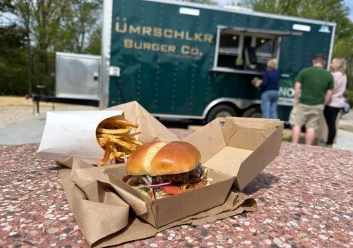 A burger and fries in takeout containers, with a food truck in the background and people ordering.