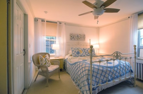 Cozy bedroom with a white metal bed, wicker chair, and soft lighting from lamps, featuring light-colored walls and decor.
