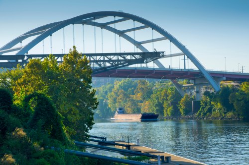 A boat navigates a river beneath a large, arched bridge surrounded by lush greenery and clear blue skies.