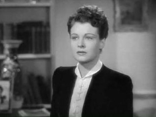 A woman with short, curly hair wearing a black jacket and white blouse, looking thoughtfully in a vintage setting.