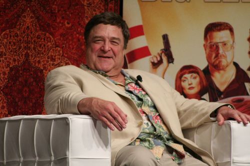 A smiling man in a light suit and floral shirt sits on a white chair, with a colorful backdrop behind him.