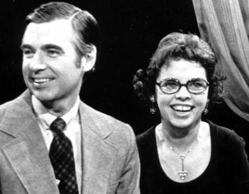 A man in a suit and a woman in glasses smile together on a television set.