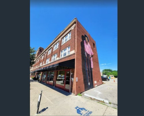 A large mural of a man in a pink shirt on the side of a brick building under a clear blue sky.