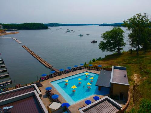 Aerial view of a pool surrounded by umbrellas, with a lake and boats in the background on a sunny day.