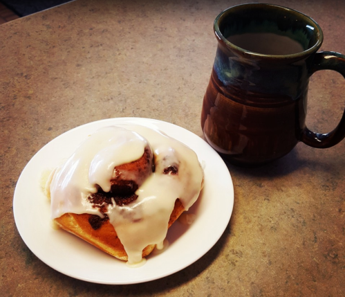 A cinnamon roll with icing on a white plate next to a brown ceramic mug filled with coffee.