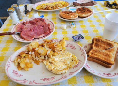 A breakfast spread featuring ham, eggs, hash browns, toast, and pastries on a checkered tablecloth.