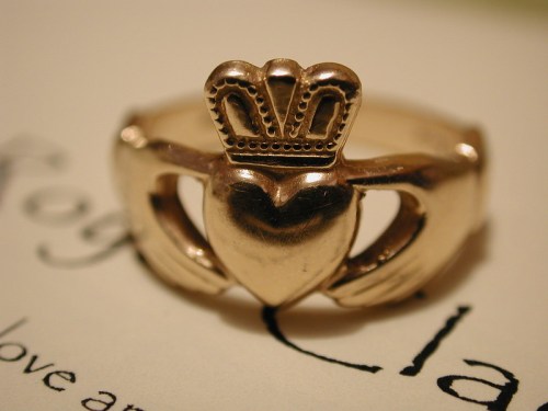 A gold ring featuring a heart held by two hands, topped with a small crown.