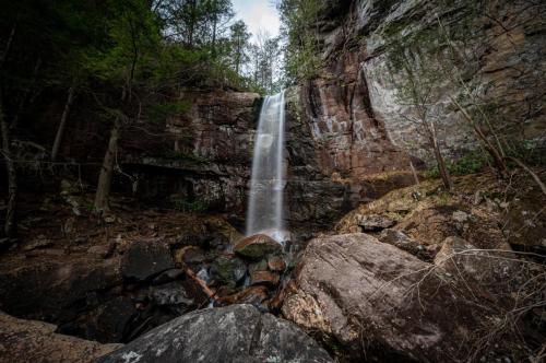 A serene waterfall cascades over rocky cliffs, surrounded by lush greenery and boulders in a tranquil forest setting.