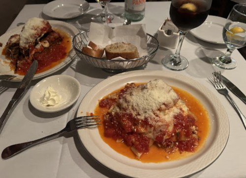 Two plates of pasta with marinara sauce and cheese, accompanied by bread, drinks, and a side of butter.