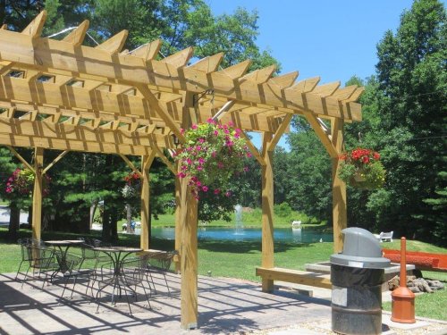 A wooden pergola with hanging flowers overlooks a grassy area and a pond, featuring a picnic table and grill.