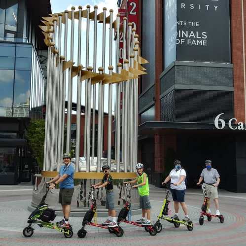 A group of five people on scooters poses in front of a large sculpture outside a sports venue.