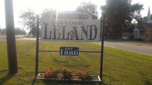 Sign welcoming visitors to Historic Leland, established in 1886, with flowers and trees in the background.