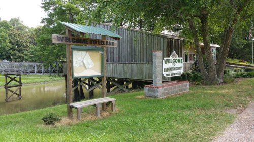 Welcome signs for Leland and Brunswick County near a wooden building by a calm waterway, surrounded by greenery.