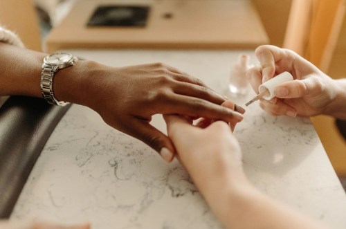 A close-up of two hands at a nail salon, one receiving a manicure with nail polish being applied.