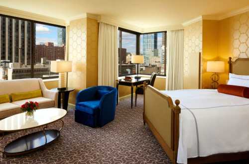 A stylish hotel room featuring a bed, seating area, desk, and large windows with city views.