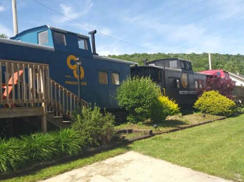 A blue train caboose and a black locomotive are displayed alongside a wooden deck and lush greenery under a clear sky.
