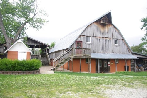 A rustic barn with a sloped roof, wooden siding, and a staircase, surrounded by greenery and a gravel path.