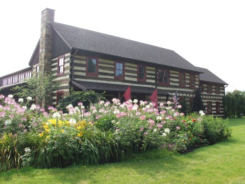 A large log cabin-style house surrounded by colorful flower gardens and greenery on a sunny day.
