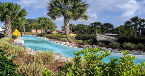 A serene waterway surrounded by palm trees and lush greenery, with a boat and colorful tents in the background.